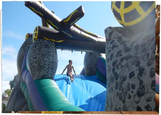 Inflatable water slide. Boy is at the top of the slide. Pillars resemble rocks and roof, logs.